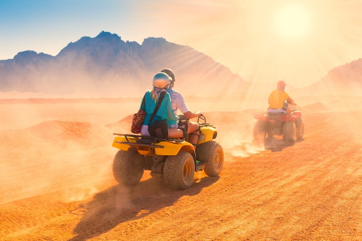 Trois pilotes en quad traversent une plaine désertique poussiéreuse du Sinaï au soleil rasant, montagnes sombres et déchiquetées à l'horizon, poussière derrière les roues.